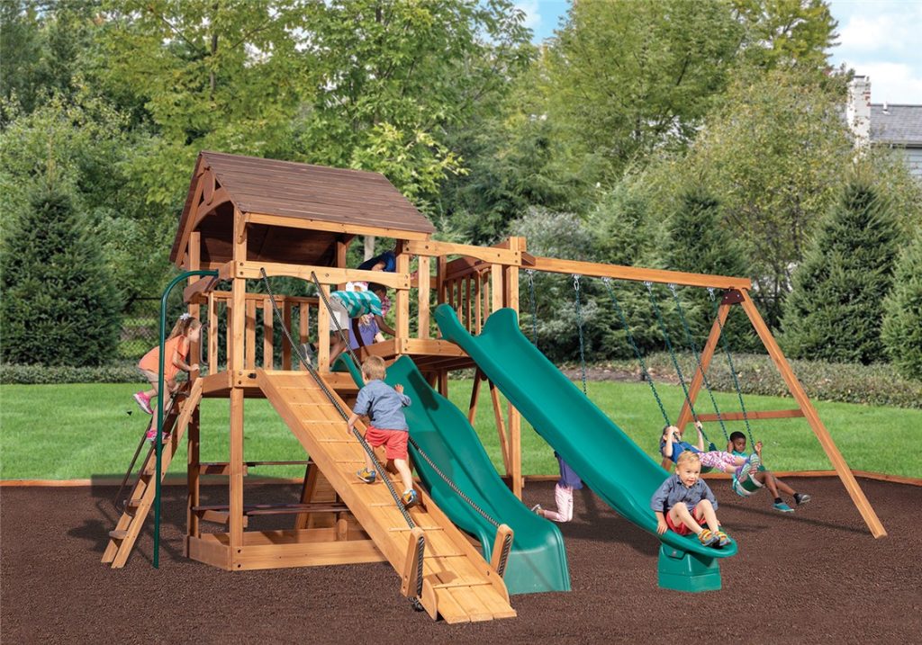 A vibrant scene of a wooden playset in a lush green backyard, with several children actively playing. The playset features a main tower with a brown roof, multiple slides including a green wavy slide and a wooden slide, and a swing set. Children are seen climbing ladders, sliding down, and swinging. The ground is covered with dark brown mulch, surrounded by green grass and abundant trees in the background.