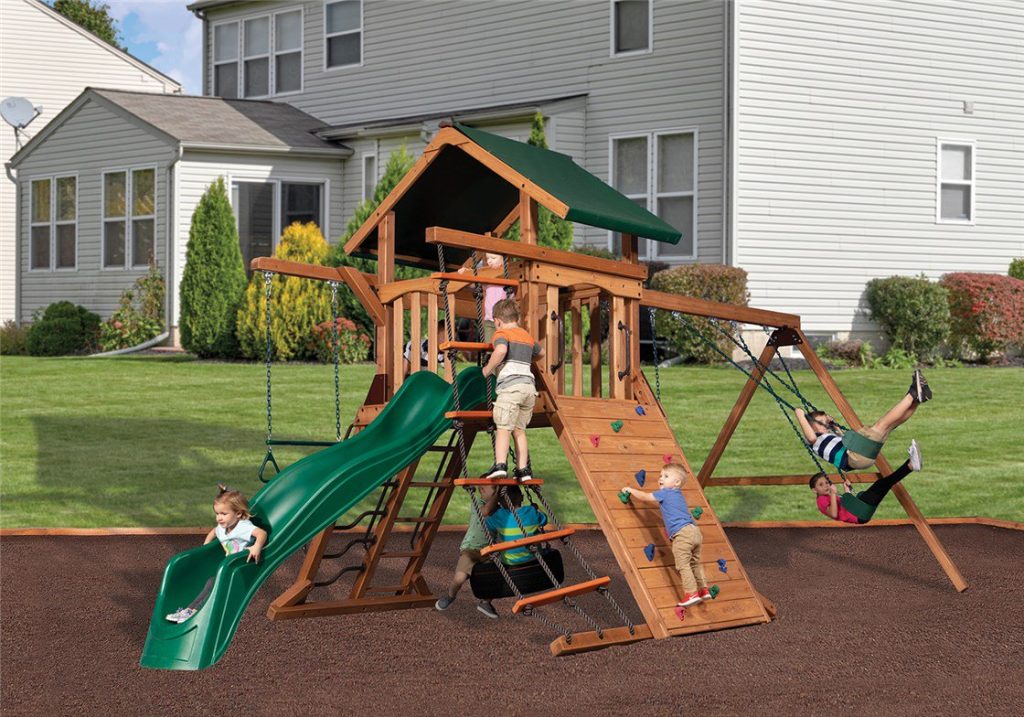A large wooden playset with a green slide and roof is bustling with children playing in a backyard. A young girl in a white shirt is at the bottom of the green slide, while a boy in an orange shirt is climbing up the playset's ladder. Another child is climbing a rock wall, and two more children are on swings. The playset is situated on a dark brown surface, with green grass and a house in the background under a partly cloudy sky.