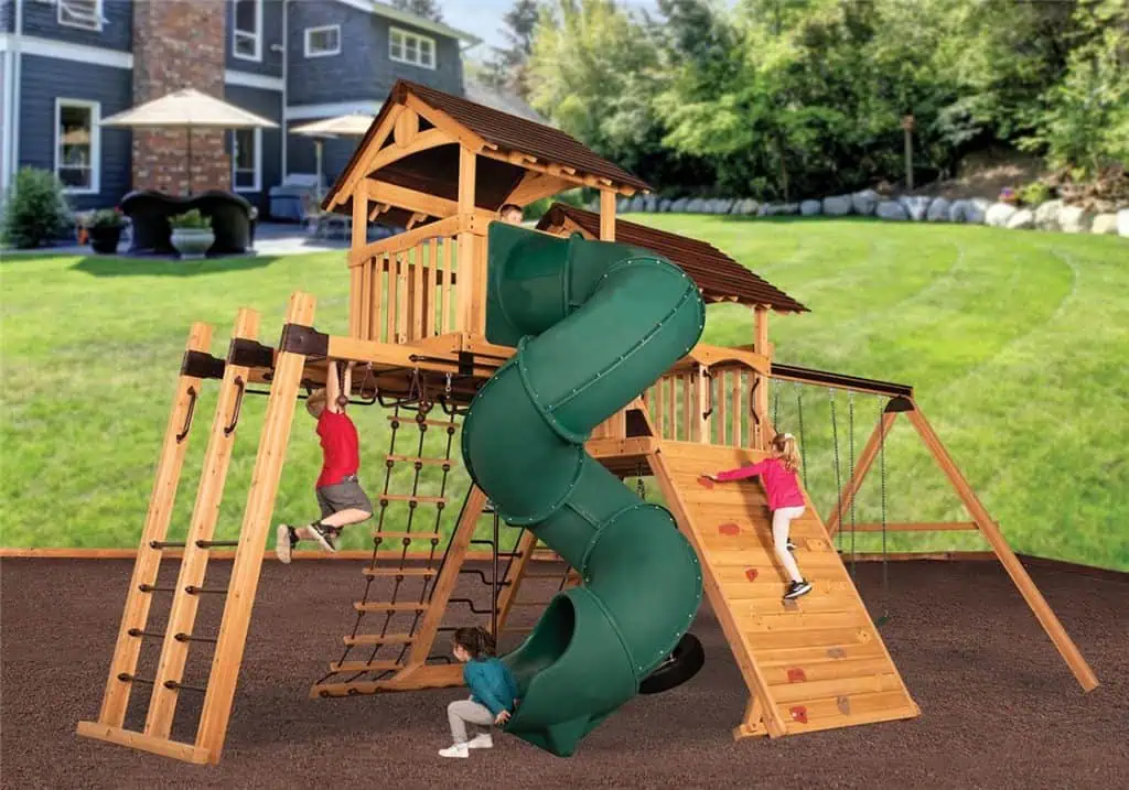 A vibrant wooden playground, featuring a large green spiral slide, is filled with children engaged in various activities. A girl in a pink top climbs a wooden rock wall on the right, while another child sits at the bottom of the spiral slide. A boy in a red shirt hangs from monkey bars on the left. The playset has multiple climbing features, swings, and a tower with a brown roof. It's set on a dark brown surface in a lush green backyard, with a large house visible in the background under a bright sky.